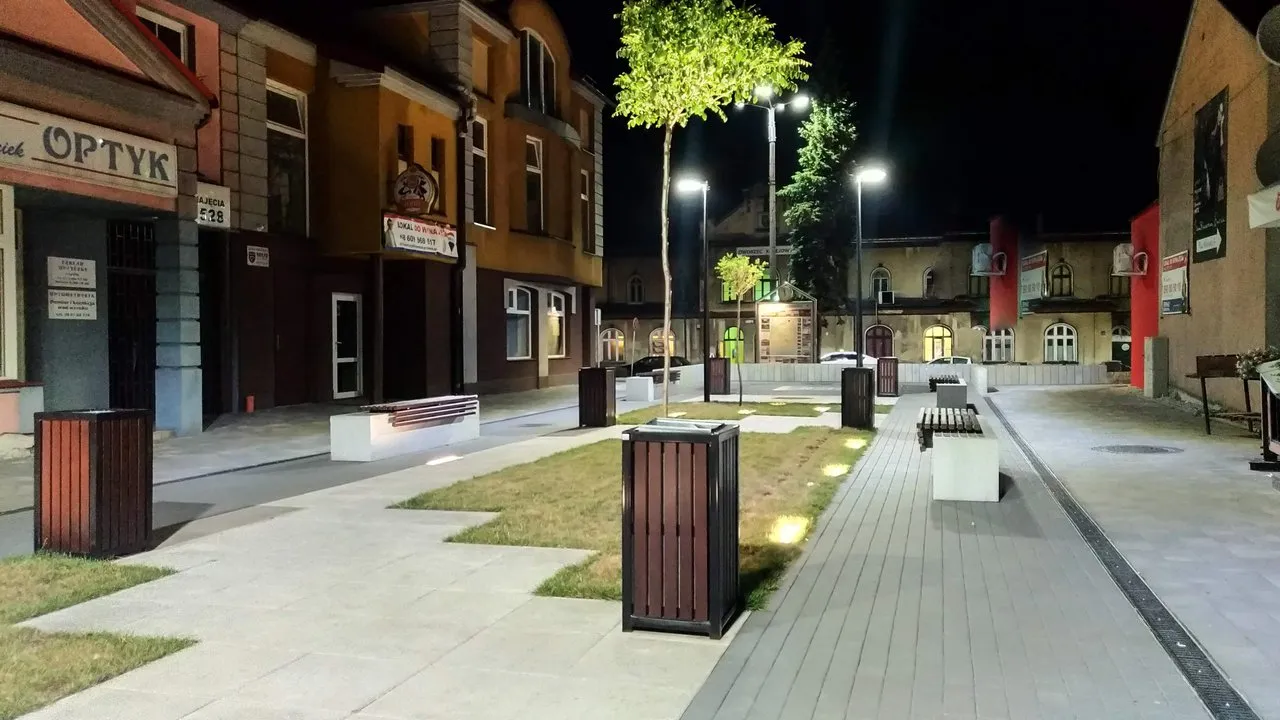 A night photography of a yard in front of restaurants in Polish town. Few benches and trees in the middle of the image and a railway station in the background.